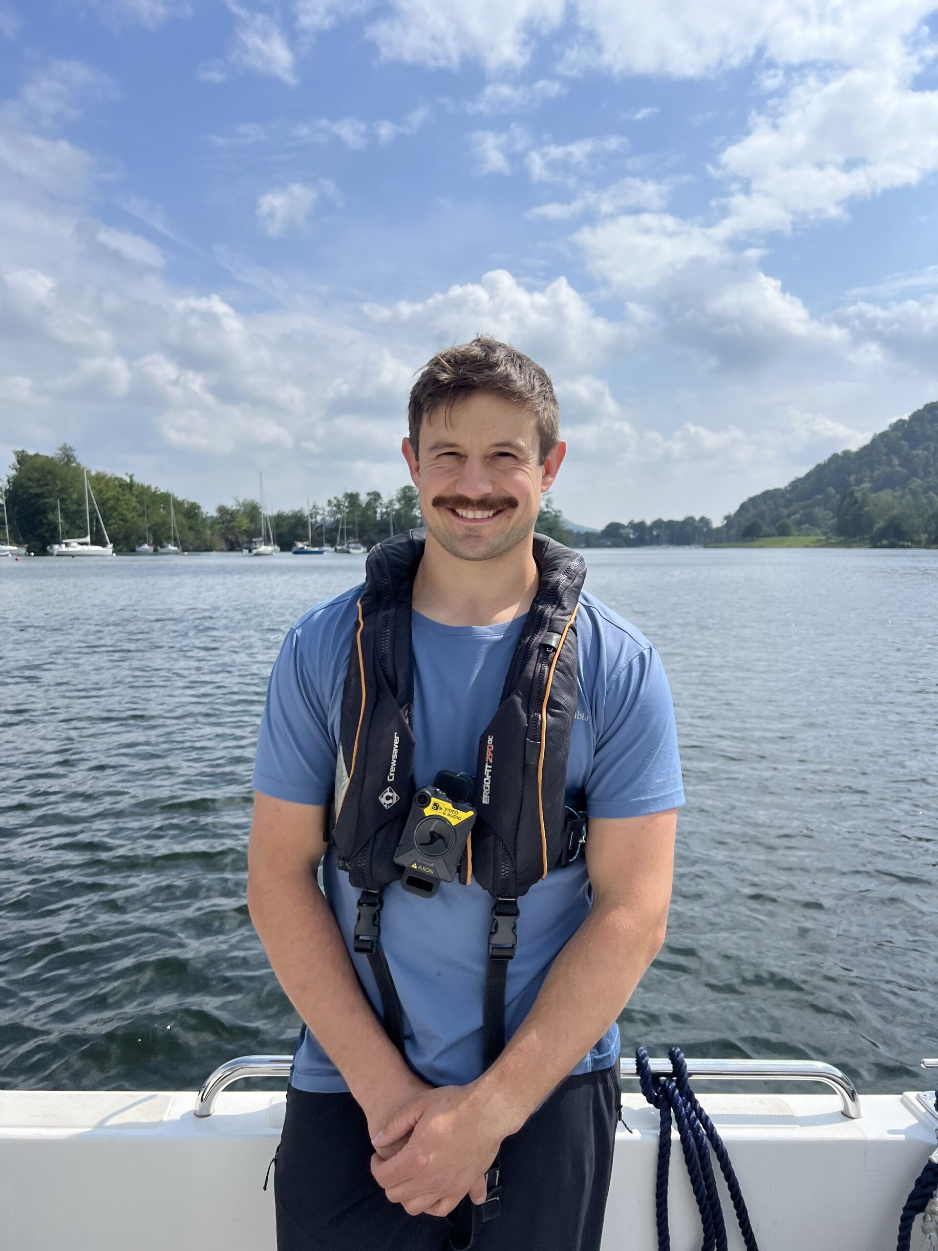 A man on a boat smiling at the camera with water, boats and a blue sky in the background