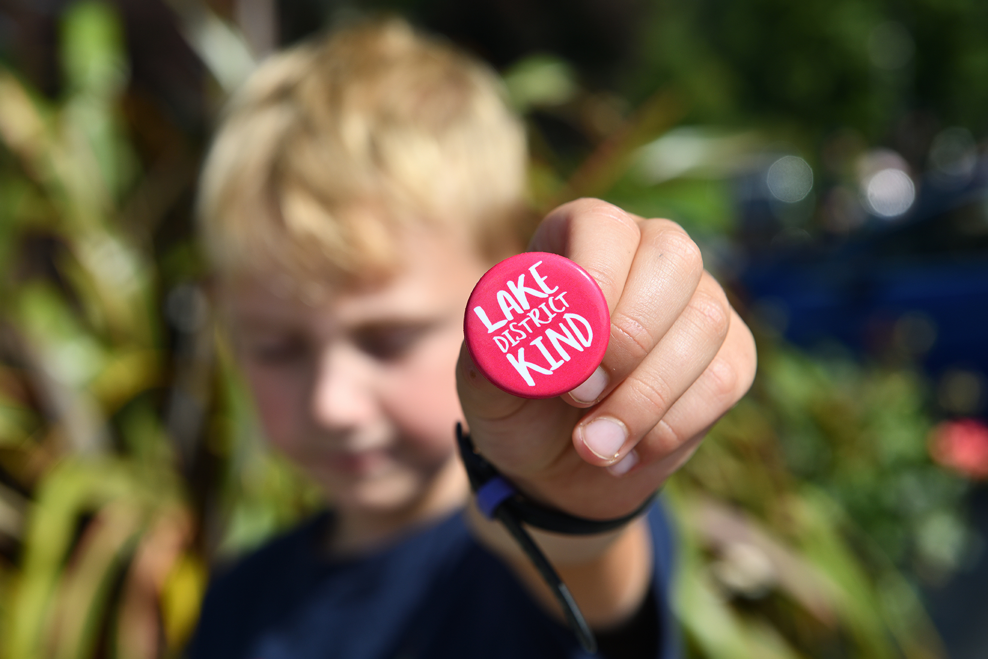 Out of focus young boy holding pink Lake District Kind pin badge to camera