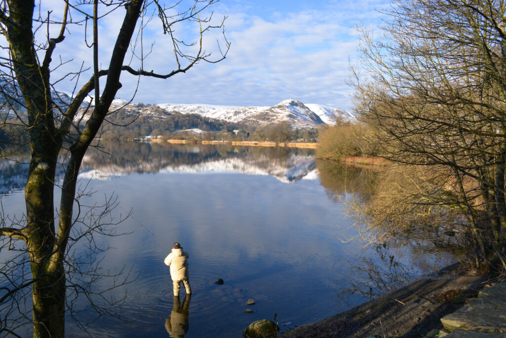 Man stood in still lake with fishing rod with snowy-topped mountains in the distance