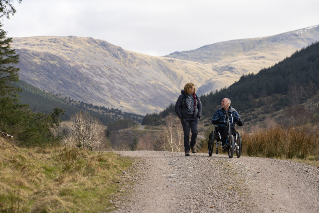 women walking alongside a man in a wheelchair bike going along a gravelled path with mighty fells in the distance