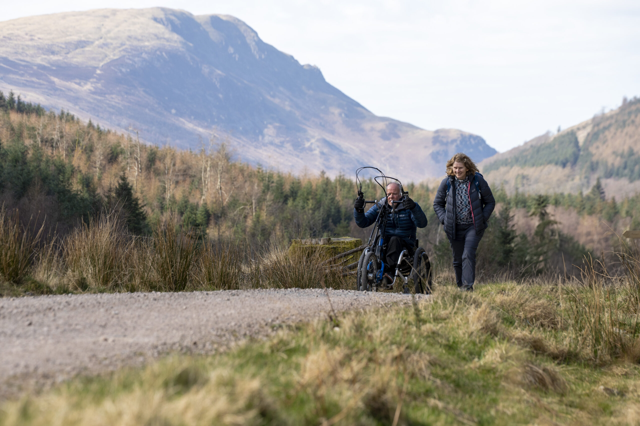 white adult female walking next to white adult male wheelchair user. On wide gravel path, surrounded by grey and green woodland and fells
