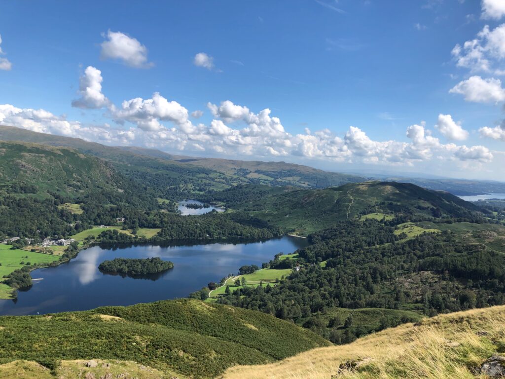 view of derwent water and surrounding green fields, woodland and fells, blue sky with a couple of white clouds
