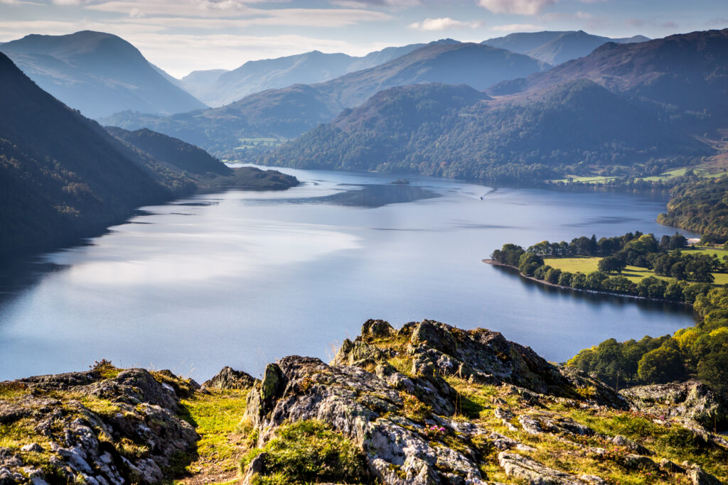 Arial view of Ullswater, large light blue lake body surrounded by green woodland, fields and brown fells