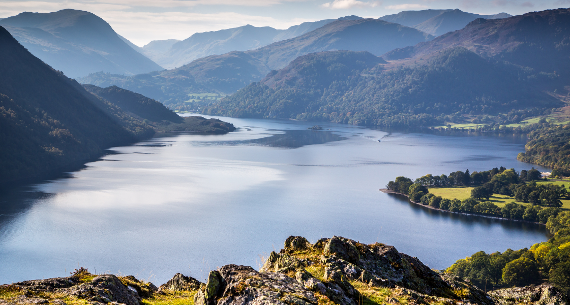 Arial view of Ullswater, large light blue lake body surrounded by green woodland, fields and brown fells