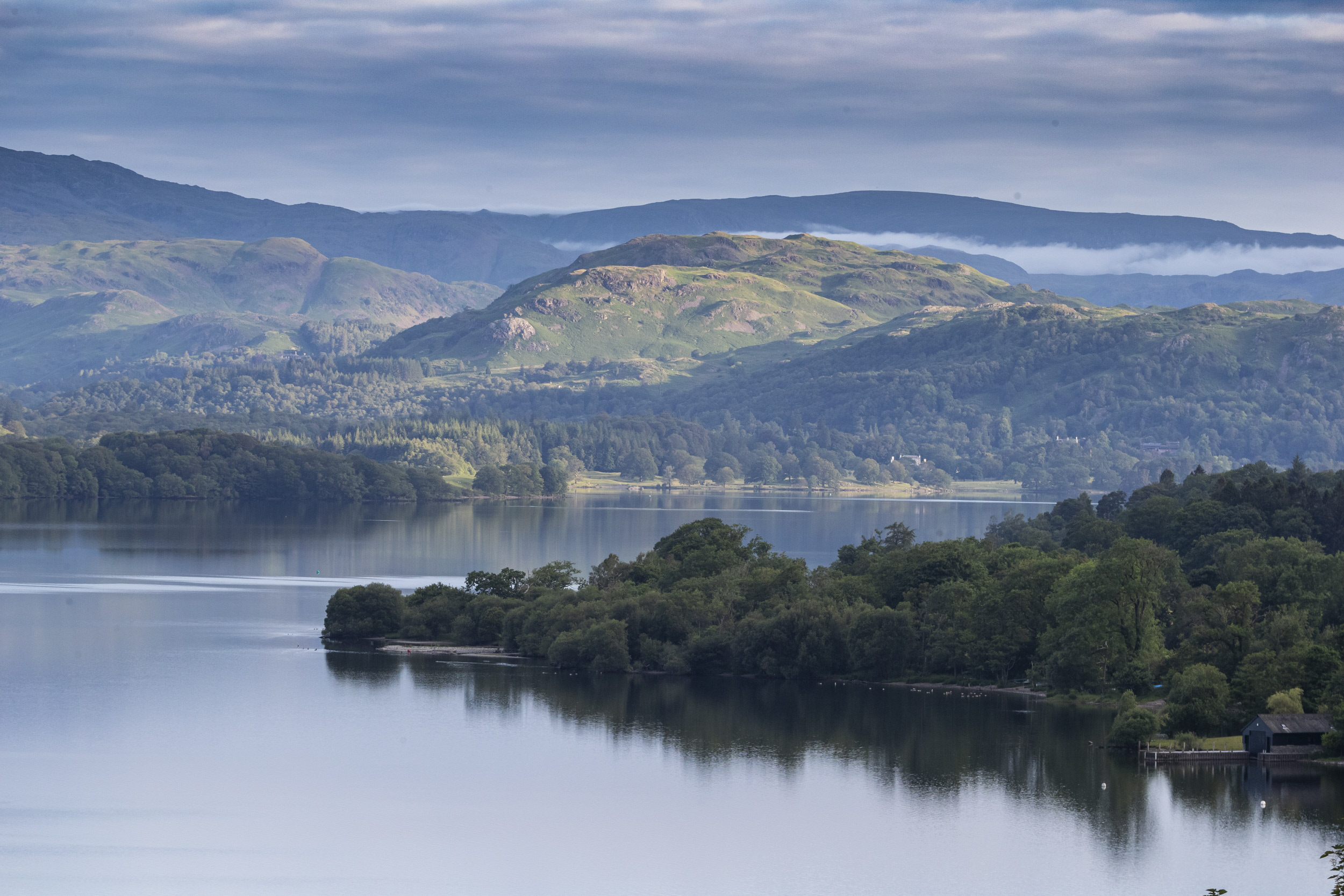 Landscape view of Windermere. Blue and grey lake and sky, green woodland extending into lake, green fells in background