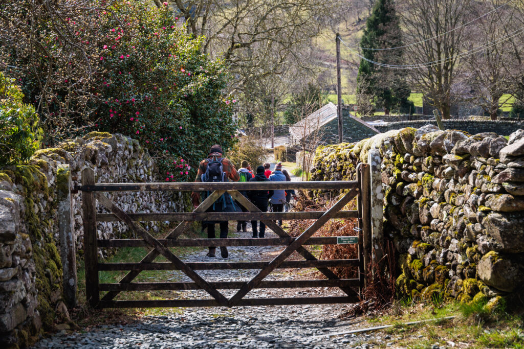 Group of adult walkers on the Coast to Coast path. Wide stone path inside dry stone walls, surrounded by farm and vegetation. Walkers behind wooden gate with sign reading please close the gate behind you