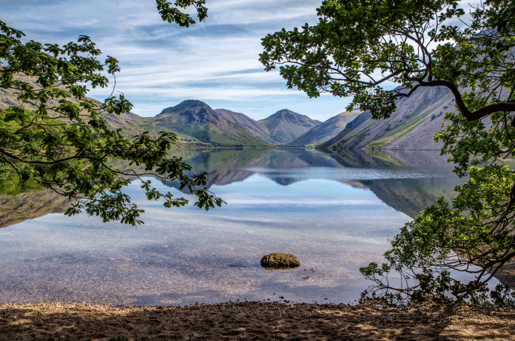 Panoramic landscape view overlooking Wastwater towards Great Gable and surrounding grey fells. Lake and fells framed by the stone lake shore foreground and overhanging tree branches with green leaves