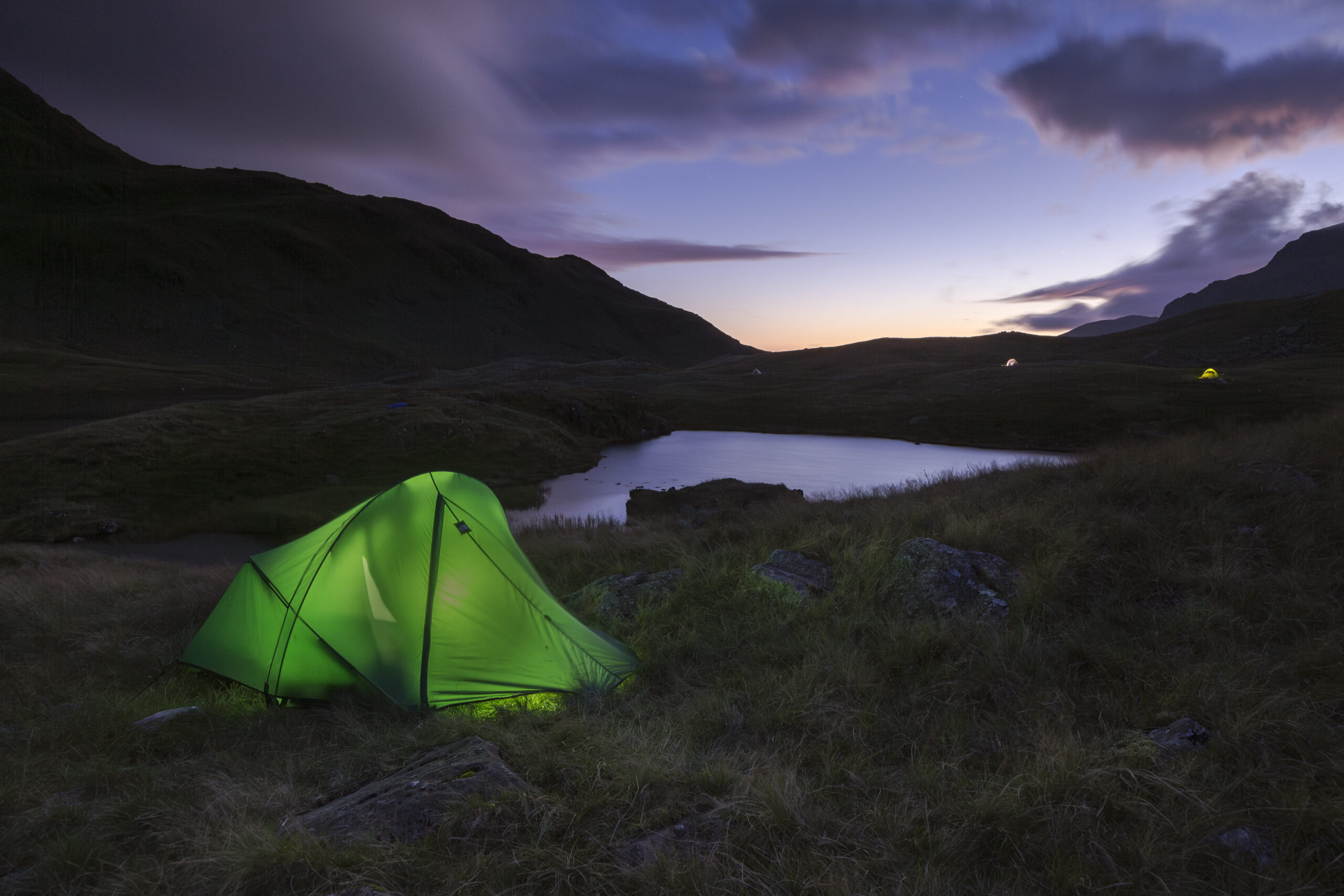 Night time landscape view of Sprinkling tarn, a small body of water high in the surrounding shadowed fells with purple sky. A torch illuminated light bright tent in foreground.