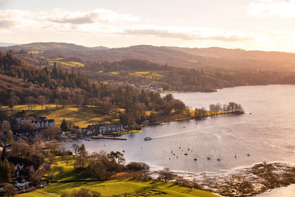 Aerial view of lake Windermere at sunset/ Purple blue lake, green woodland and fields, brown fells and buildings surrounding the lake. Motorised, sailing and small boats on the water.