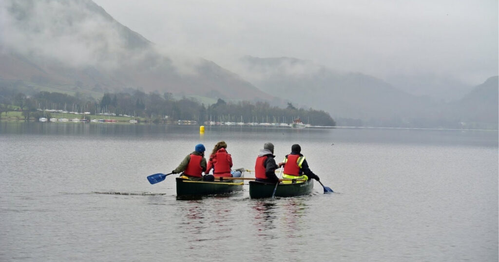 Two green canoes on the lake on a dark grey day. People sat in red buoyancy aids paddling