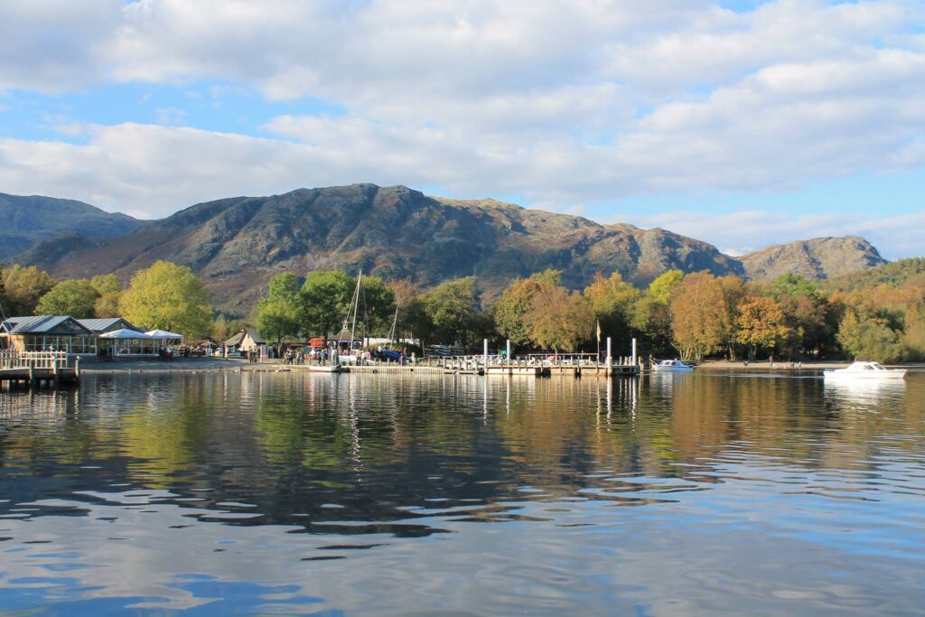 Coniston boating centre jetty and docked boats on a sunny day. Fells and trees in background, reflected in the water below