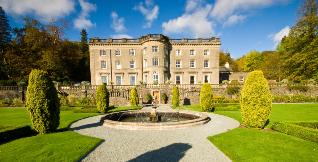 rydal hall, a grand three storey beige stone building. well kept gardens in front of the hall, with a gravel path, circular pond and fountain and small oval trees. woodland behind, blue sky day