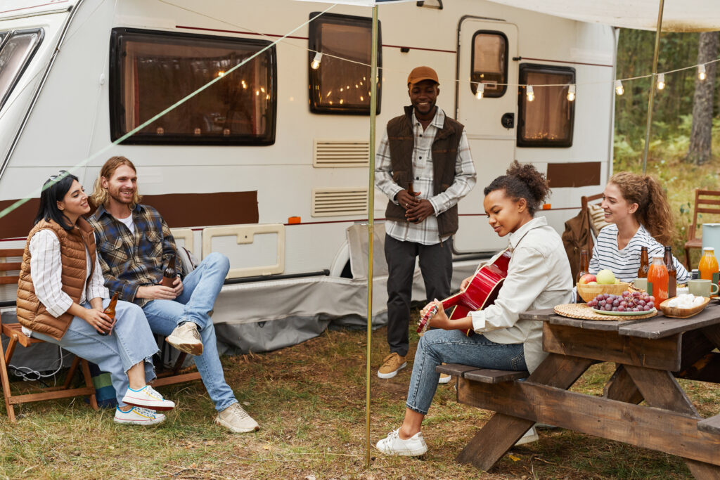 Group of five young adults smiling sat outside a caravan. Some sat on benches, one playing a red guitar under warm fairy lights. Food and drinks on one table