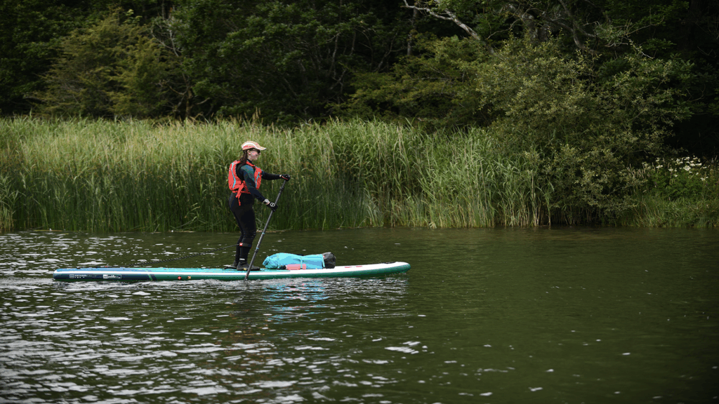 Lady on a paddleboard wearing wetsuit and buoyancy aid paddling by lakeshore