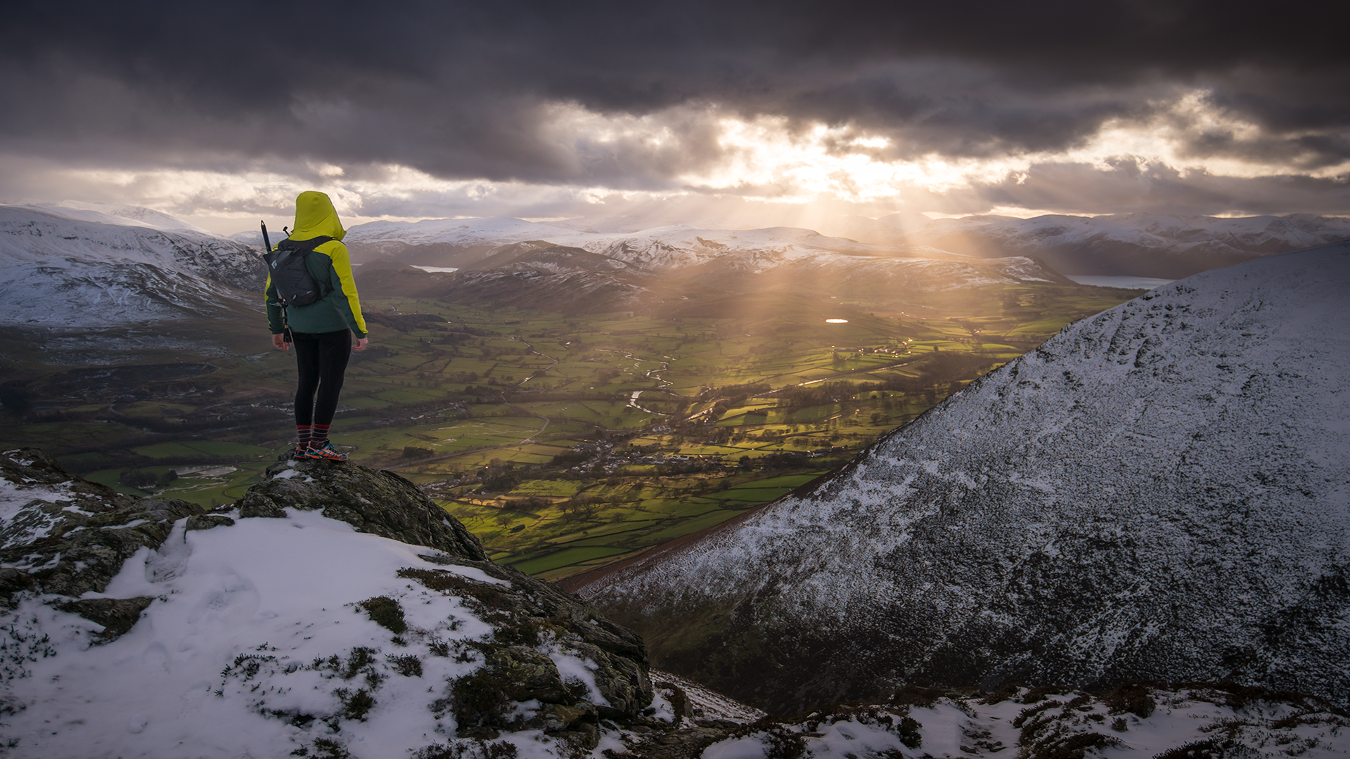 A fell walker in outdoor clothing stood on top of a snowy topped fell looking down onto the valley with sun rays coming out from the moody clouds