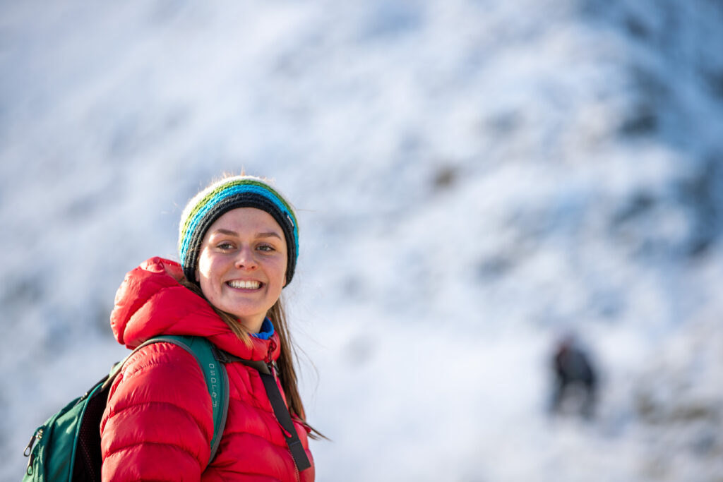 smiling white adult female in red puffer coat and blue knit hat with snow backdrop