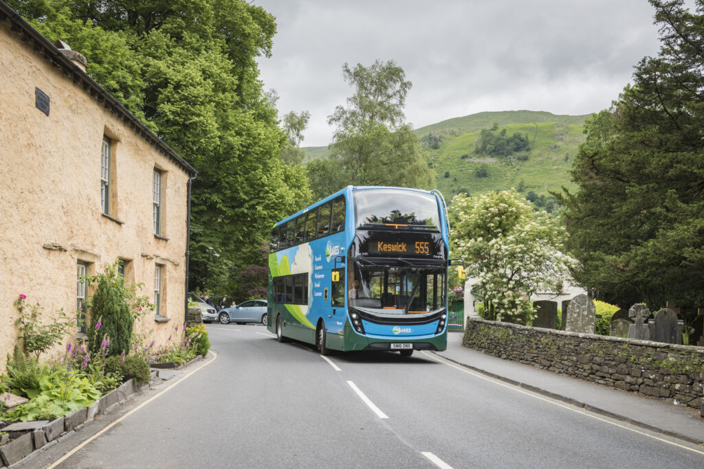 keswick 555 double decker bus driving down road with stone cottage to left and dry stone wall with blossom tree to right