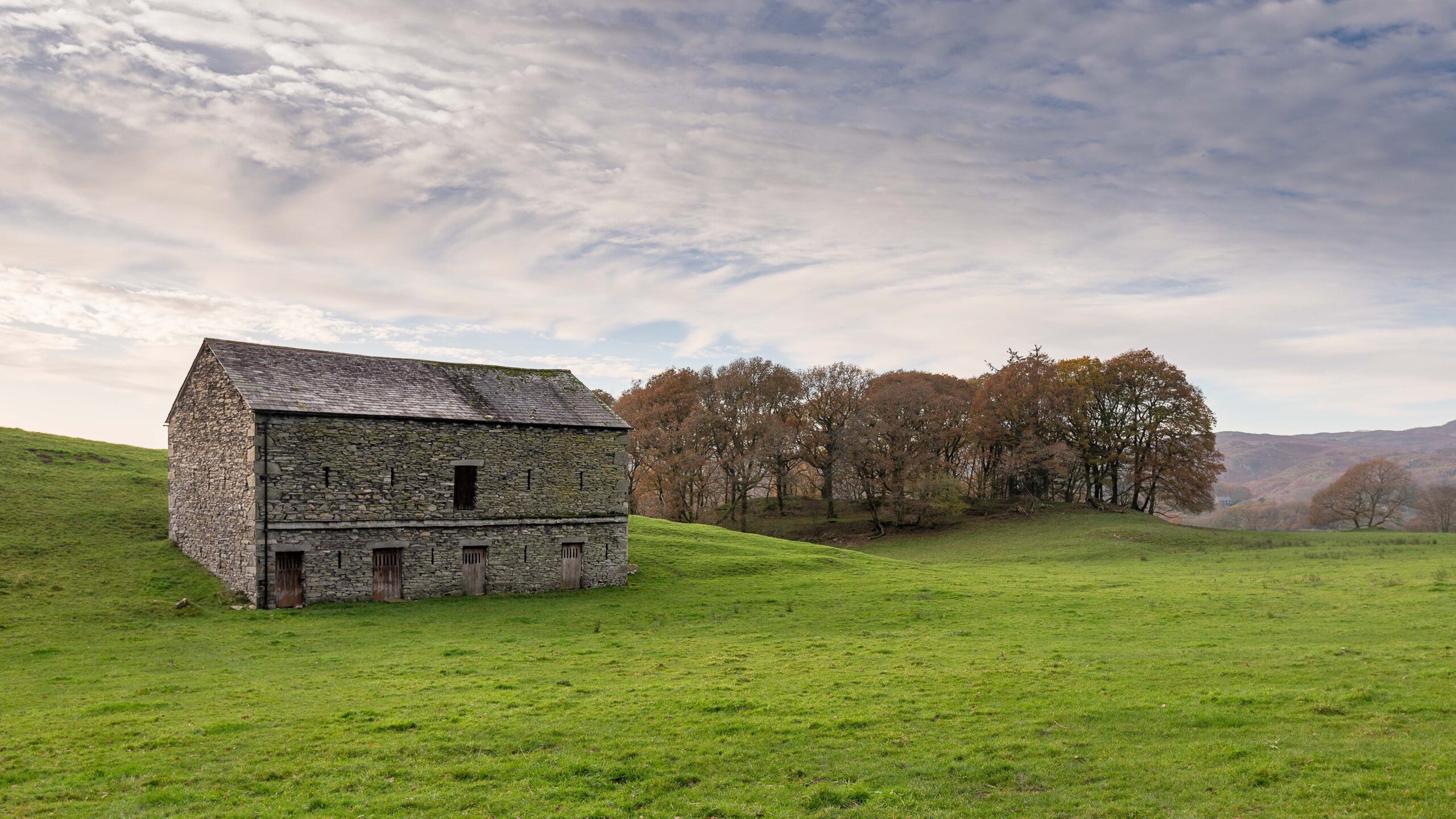 well structured stone barn on green grass field with brown patch of woodland behind