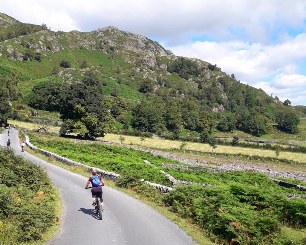 adult female cruising down tarmac lane on mountain bike. green vegetation, dry stone walls and small fell backdrop