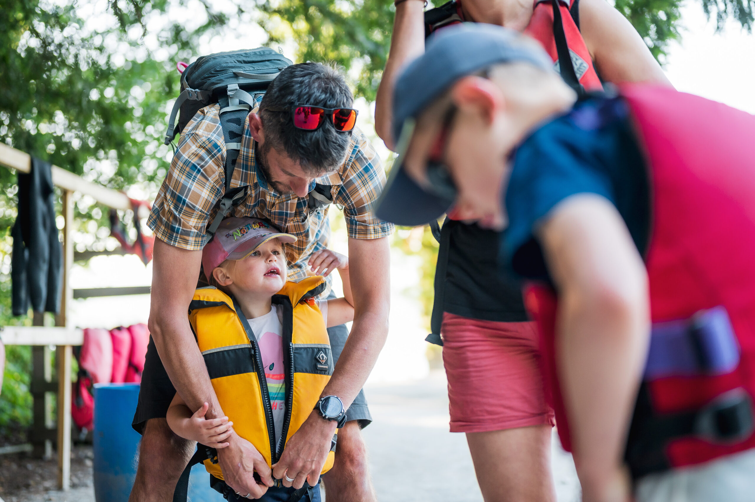 young child getting zipped into a yellow buoyancy aid by adult male, surrounded by two other adults, sunny day on the lake shore