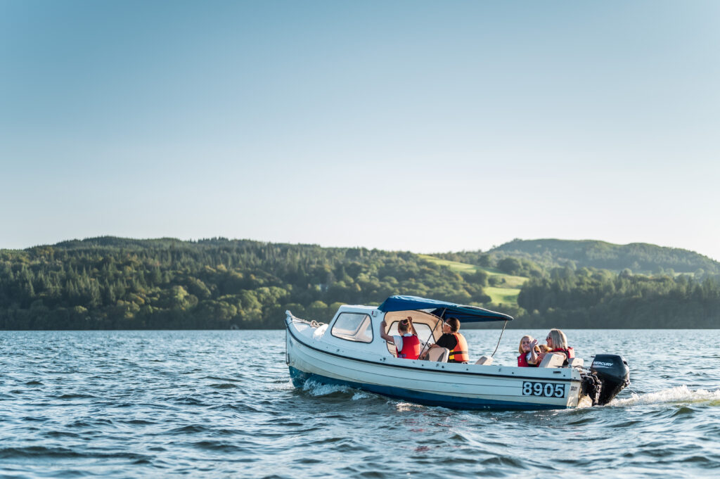 Small white motorised boat on lake, family of passengers, with fells in the background on a blue sky sunny day