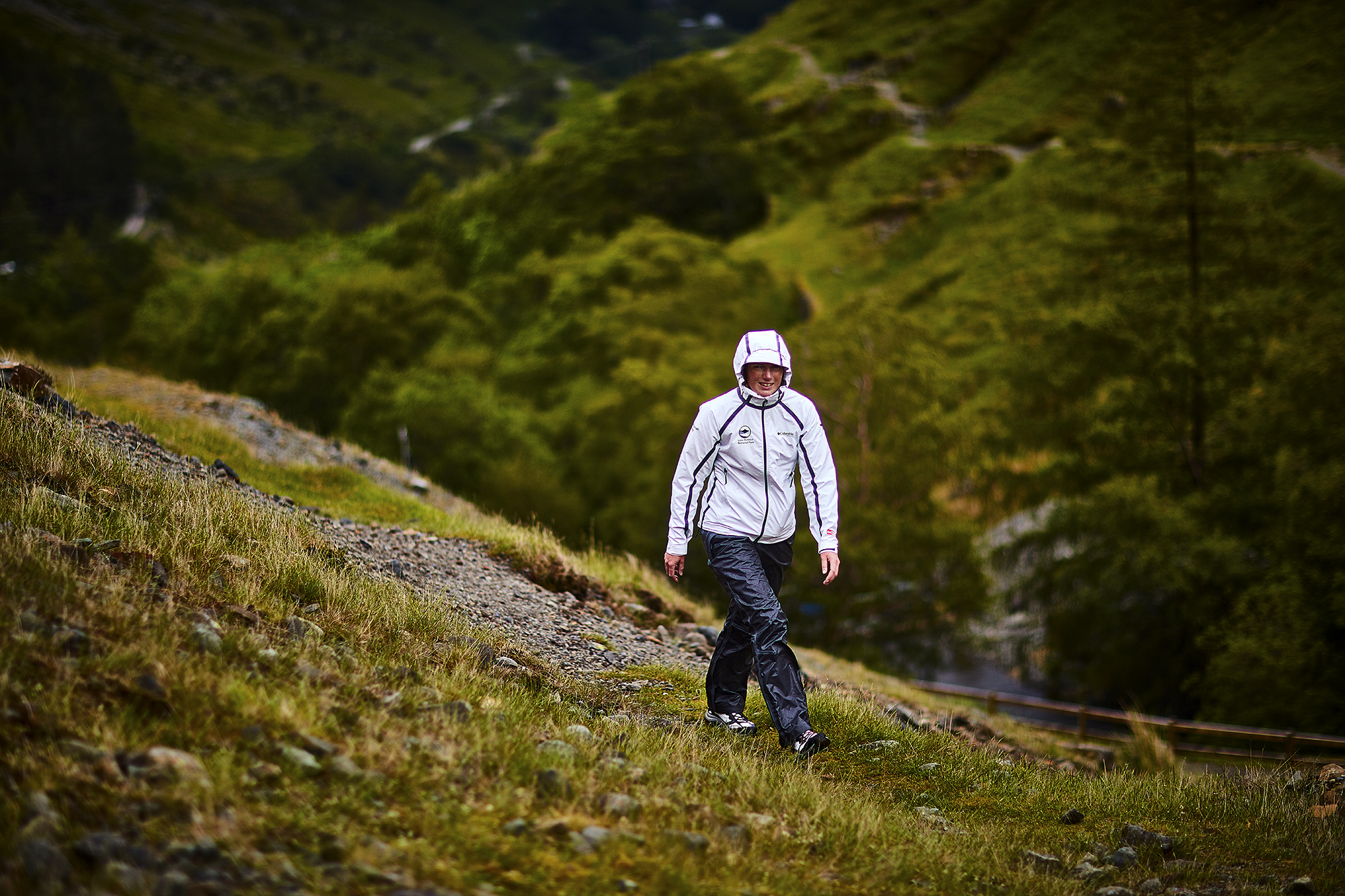 A LDNPA ranger in a white waterproof jacket walking on a hill, there are trees in the distance and flowers on the ground behind her