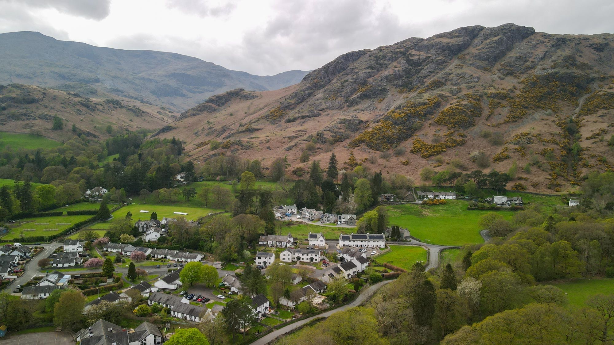 group of houses in coniston nestled at bottom of fells amongst green woodland and fields