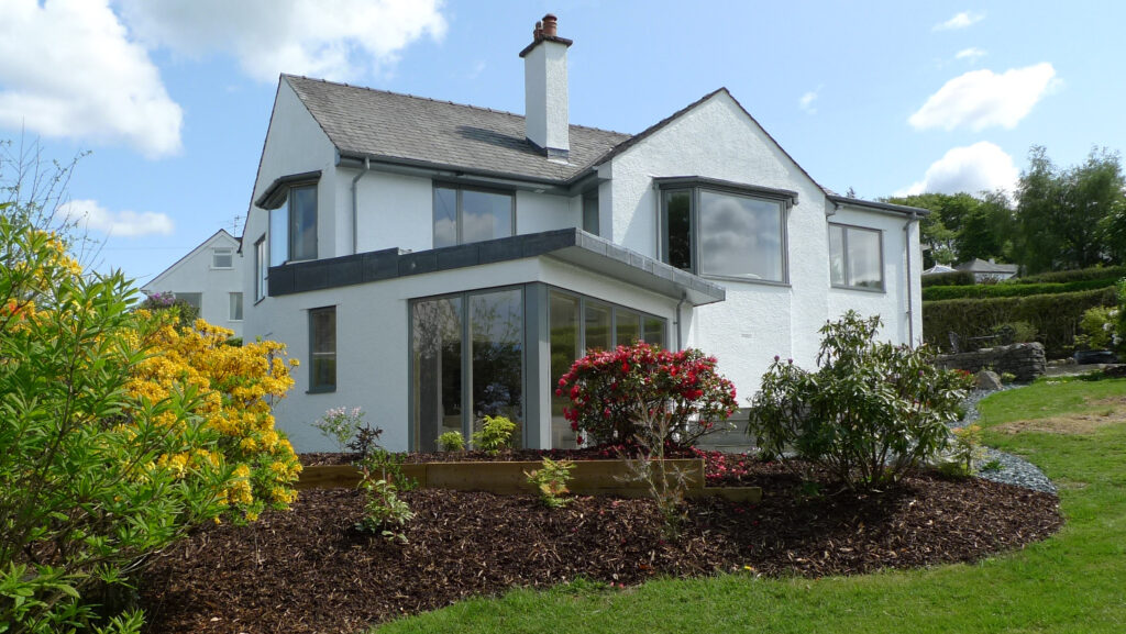 large modern white detached house with big glass windows surrounded by well kept grass garden with shrubs in flower beds
