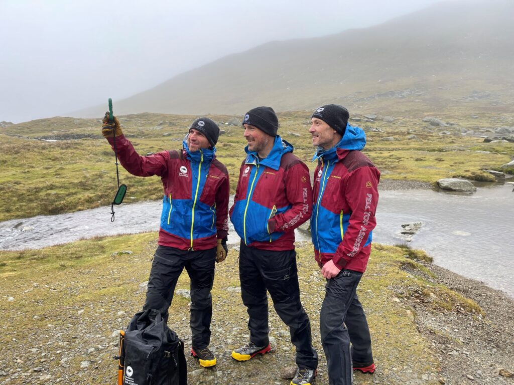 three white adult males in waterproof outdoor clothing stood together, taking a wind speed reading using a hand held device. behind them is a slightly misty, moody fell landscape and a small stream
