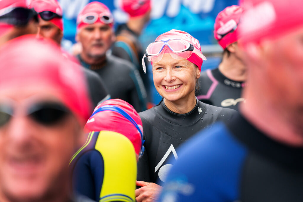 focus on adult woman smiling in wetsuit, pink swim cap and clear goggles, surrounded by more adult swimmers
