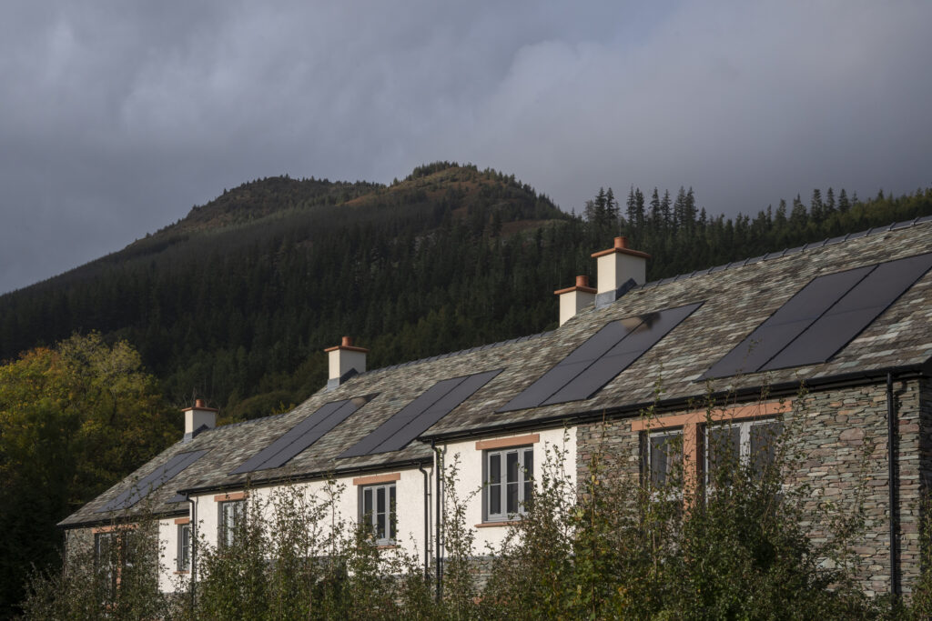 row of four slate and white houses, with slate roofs and solar panels. Only the top of the houses are visible, obstructed by small green trees. Shadowed fells in backdrop