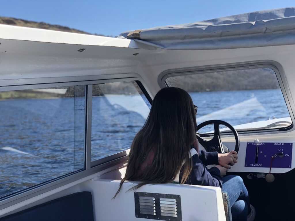 inside a small white motorised boat with adult female sat steering the wheel. blue sky and blue lake