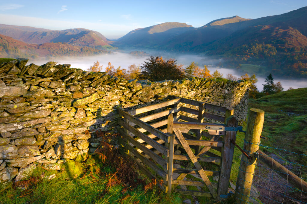 wooden kissing gate through dry stone wall with lake and fell backdrop on a warm sunset evening