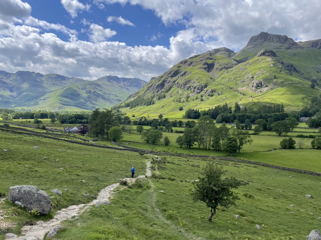 A Lake District scene with a path from the bottom left meandering into the middle of the frame and distance - a person is visible. On the horizon are Lake District fells, and the sky is blue and cloudy