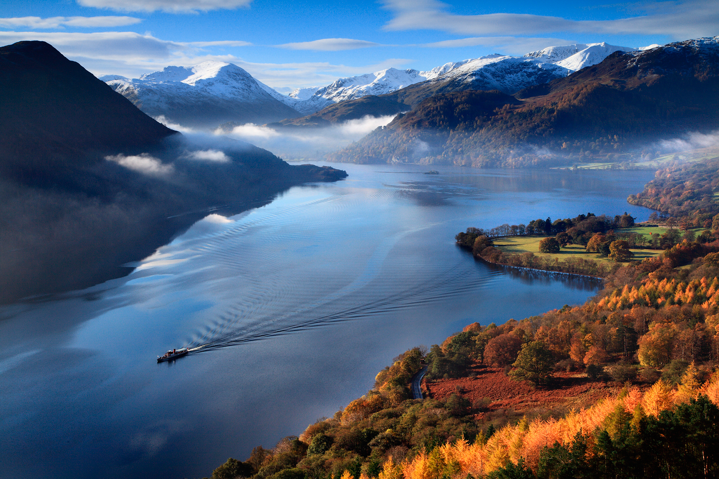 high up landscape view of deep blue ullswater, surrounding snow capped fells, autumnal orange woodland and ullswater steamer