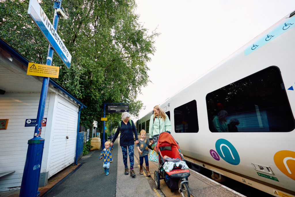 A mum pushing a pushchair with grandmother walking alongside with two young boys. Walking along a train station platform with the train stationed and sign that reads Windermere
