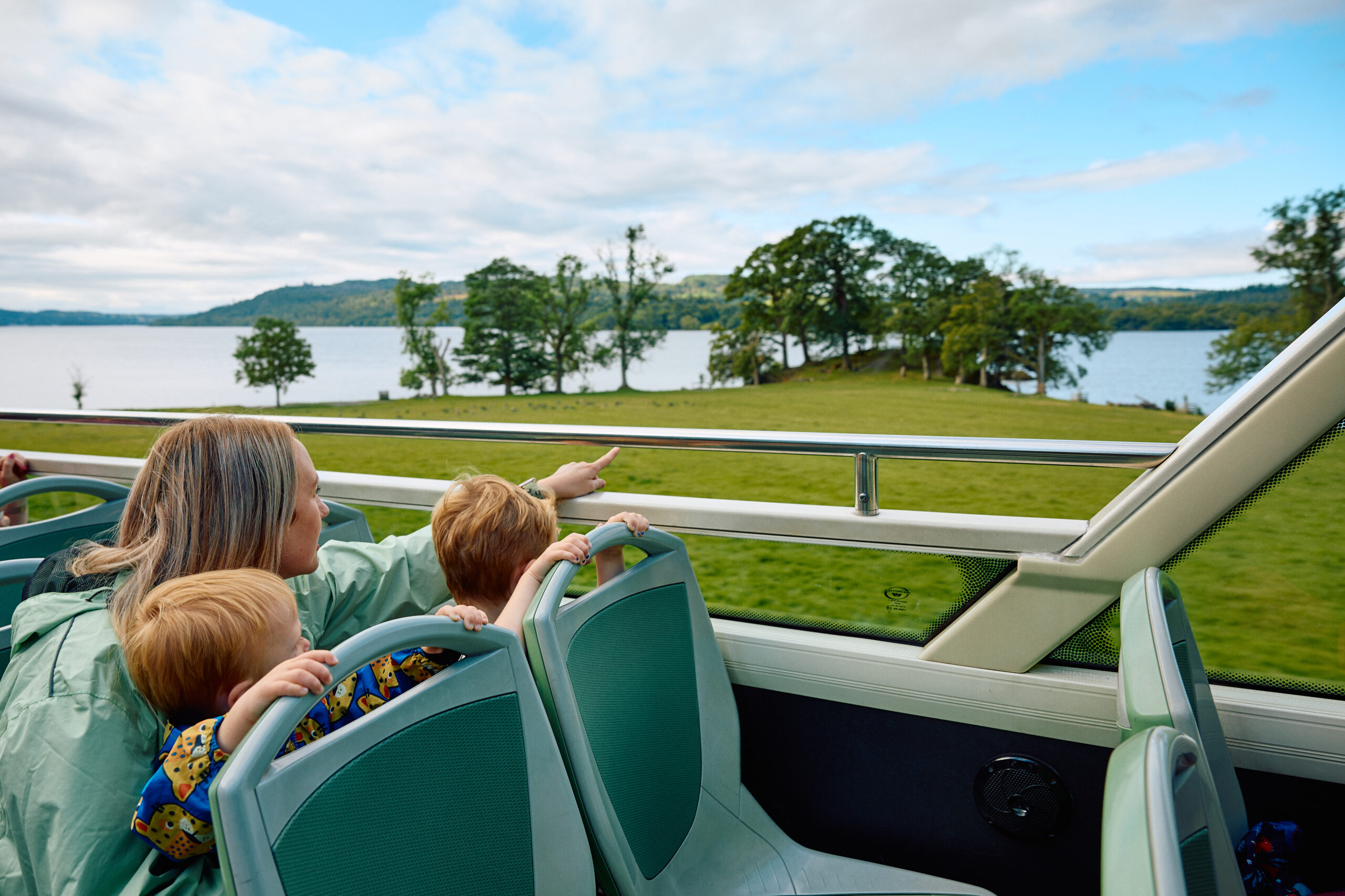 adult female and two young boys on open top bus looking and pointing out to lake and grassy tree lined lake shore