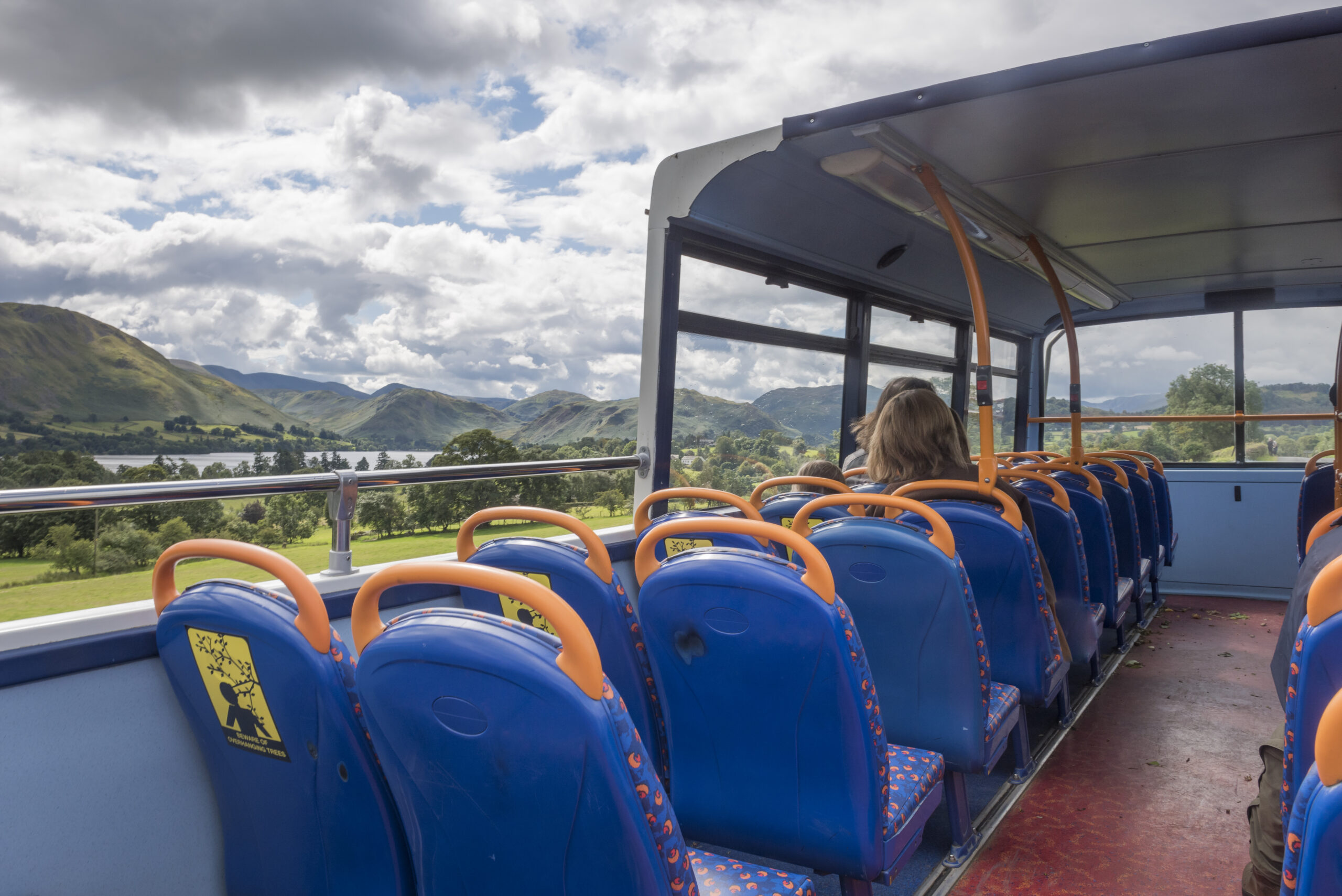 open double decker bus on a sunny cloudy day with green fells woodland and lake in distance
