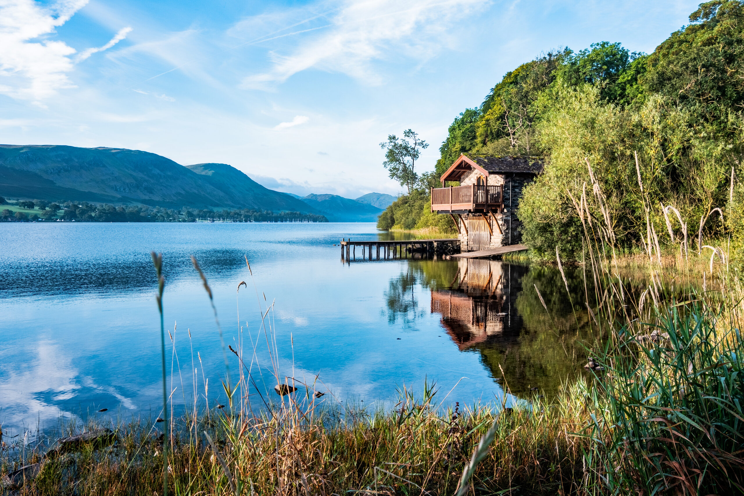 A lake on a bright blue day, on the right of the frame is a wooden boathouse with a jetty, surrounded by reeds and trees