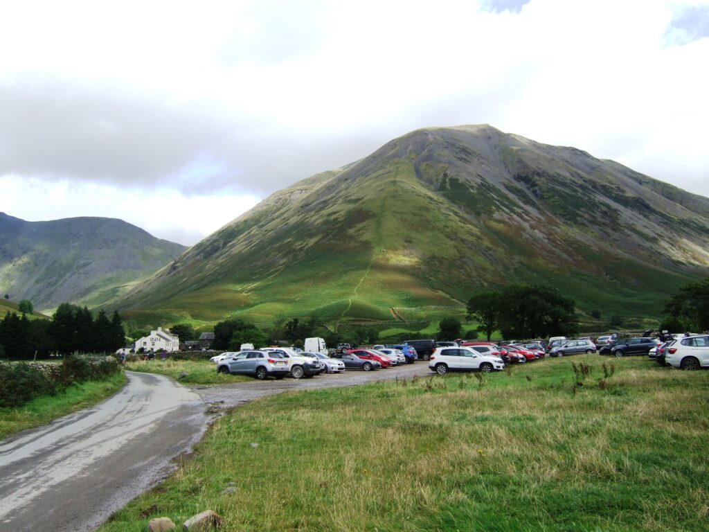 A narrow road leading to a car park full of cars at the bottom of a fell with a white building, Wasdale Inn, in the far distance
