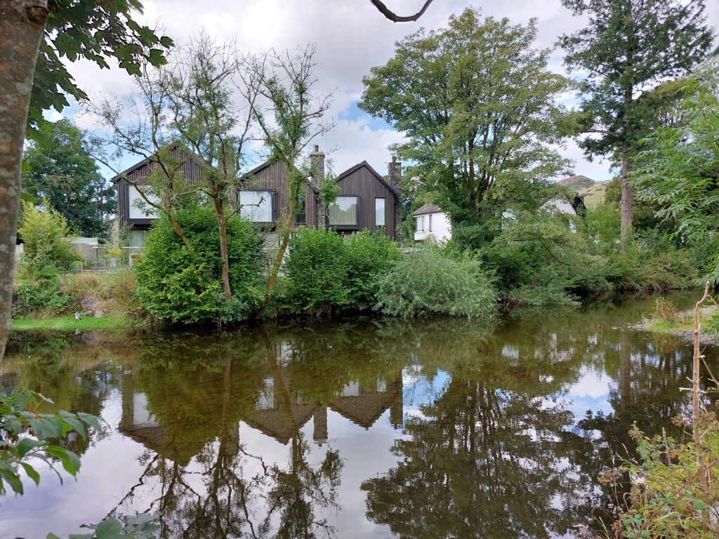 three dark wooden houses in a terrace behind bushes and trees next to a brown river