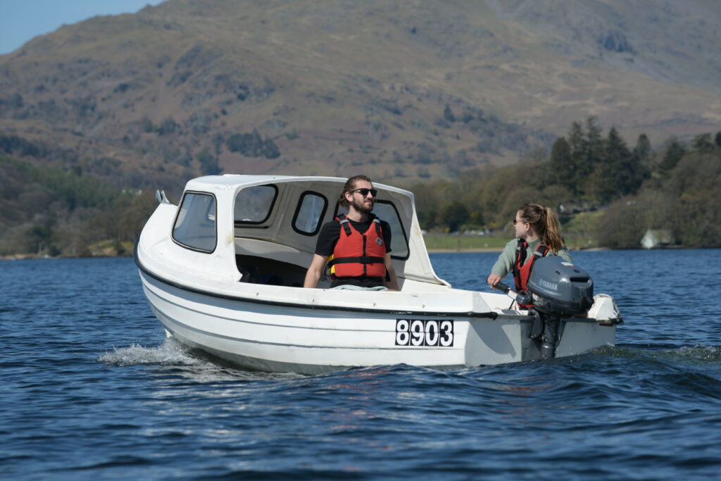 adult male and female sat in small white motorised boat on windermere with fell backdrop
