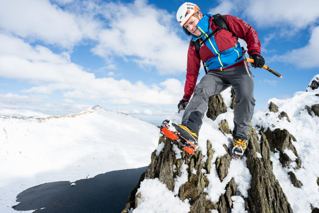 adult male in winter hiking gear with ice axe and helmet walking on steep rocky helvellyn ridge in the snow with bright sun with snow covered fell and tarn in backdrop