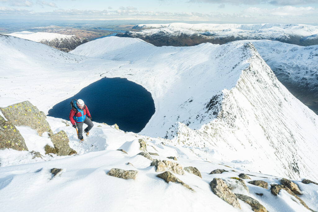 adult male in winter hiking gear with ice axe and helmet walking on steep rocky helvellyn ridge in the snow with bright sun with snow covered fell and tarn in backdrop