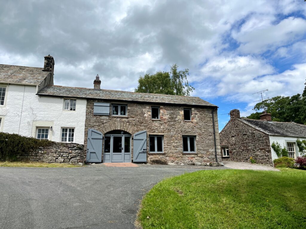 large two storey semi detached house made of large stone, with six grey framed windows and a large curved door with shutters. in front is a tarmac drive and green patch of grass, with a white house attached