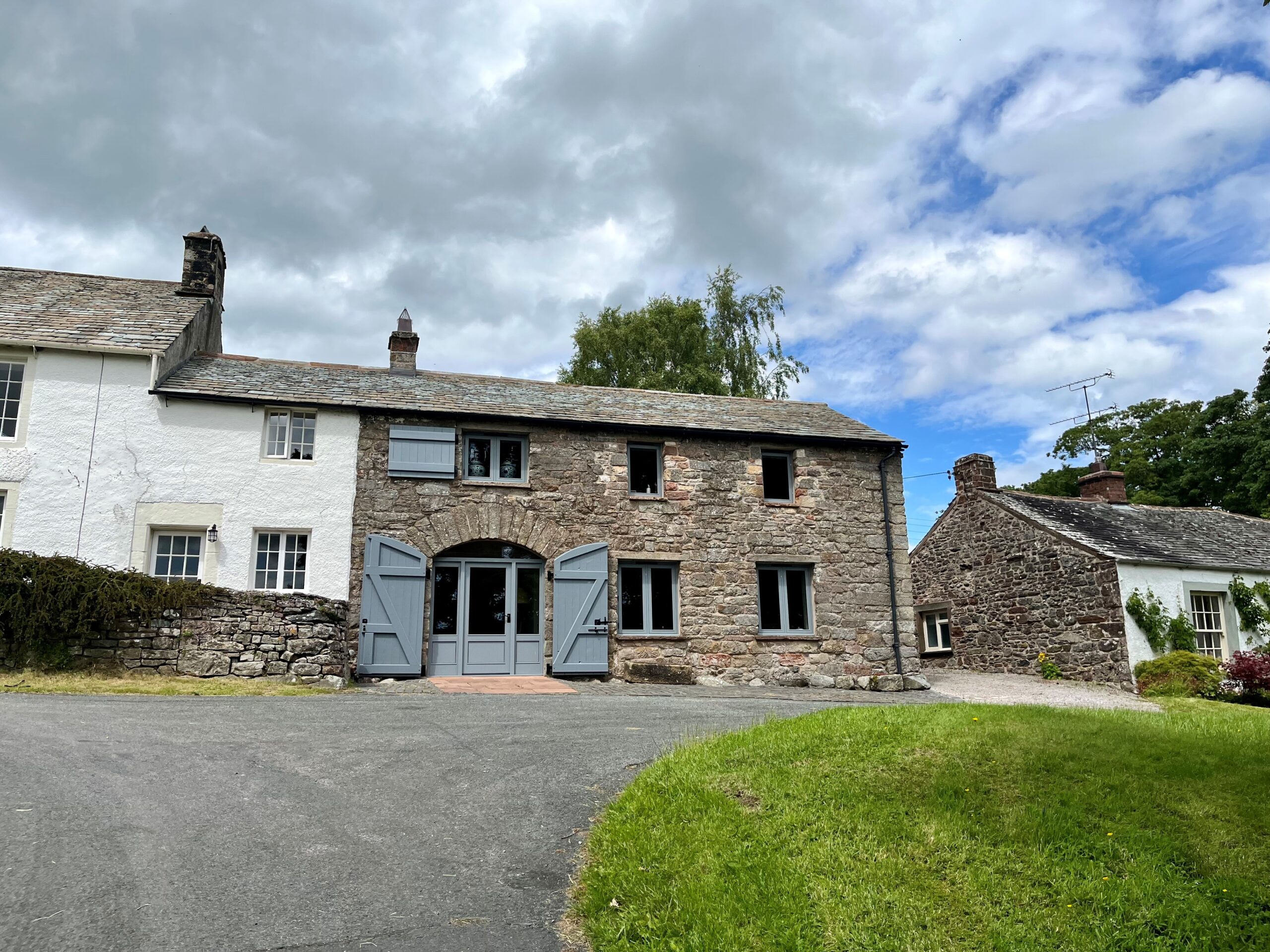 large two storey semi detached house made of large stone, with six grey framed windows and a large curved door with shutters. in front is a tarmac drive and green patch of grass, with a white house attached