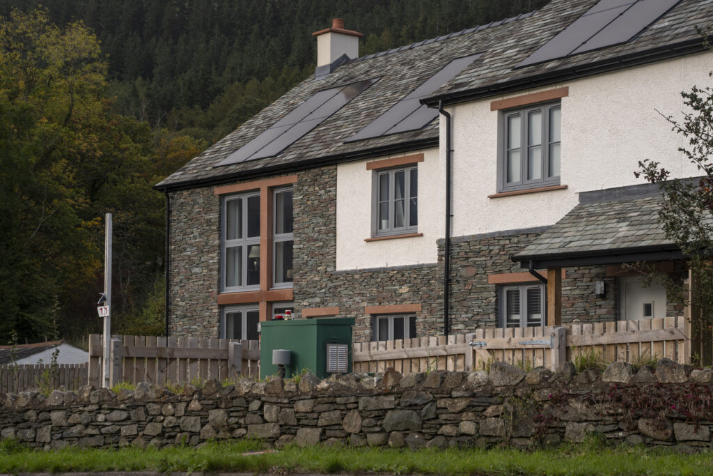 two slate and white village houses with solar panels and wooden windows behind a dry stone wall and green field