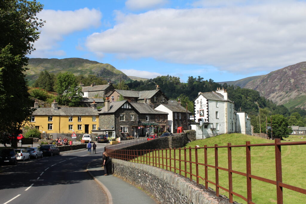 looking down a road towards a small village with a mix of orange, slate and white buildings. green field to the right of the road, divided by a stone wall and rusty metal fence. sunny day with low fells in backdrop