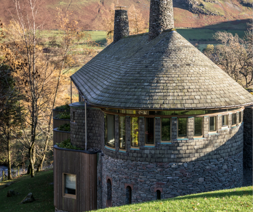 interesting architecture house by Knox Bhavan. a round, slate two storey building with a curved roof and large chimneys. thin glass rectangle windows in descending size curve round the edge of the house. thin, tiered wooden balconies with plants and a wooden extension on the ground floor. set on a grass field, with trees and grass fell bottom behind