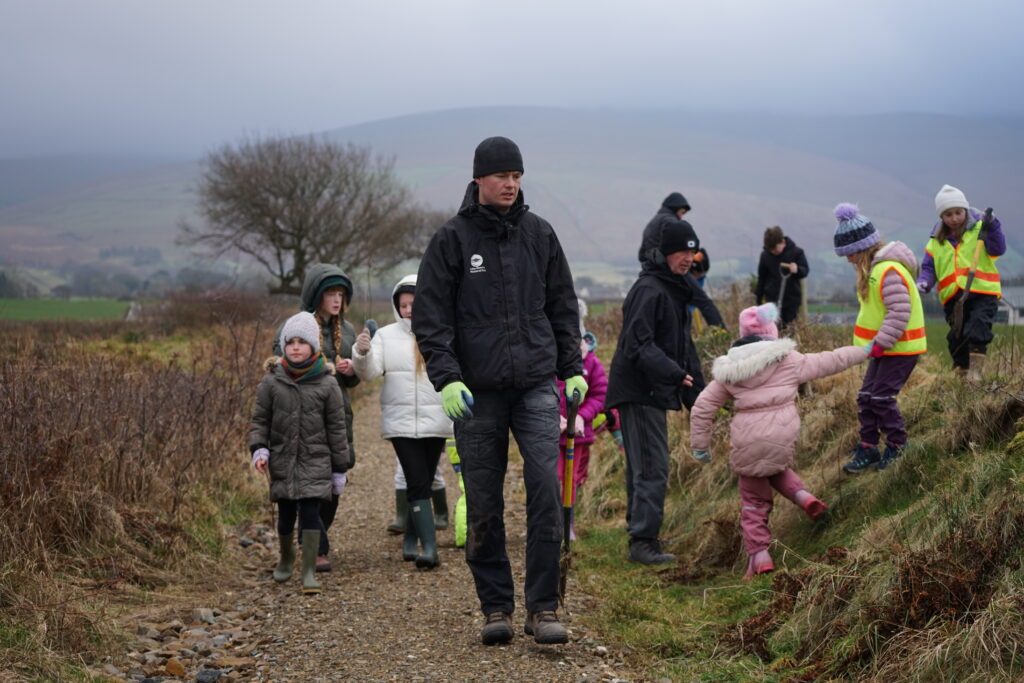 lake district national park authority ranger surrounded by children in outdoor clothing on a cloudy day in a field, planting young trees